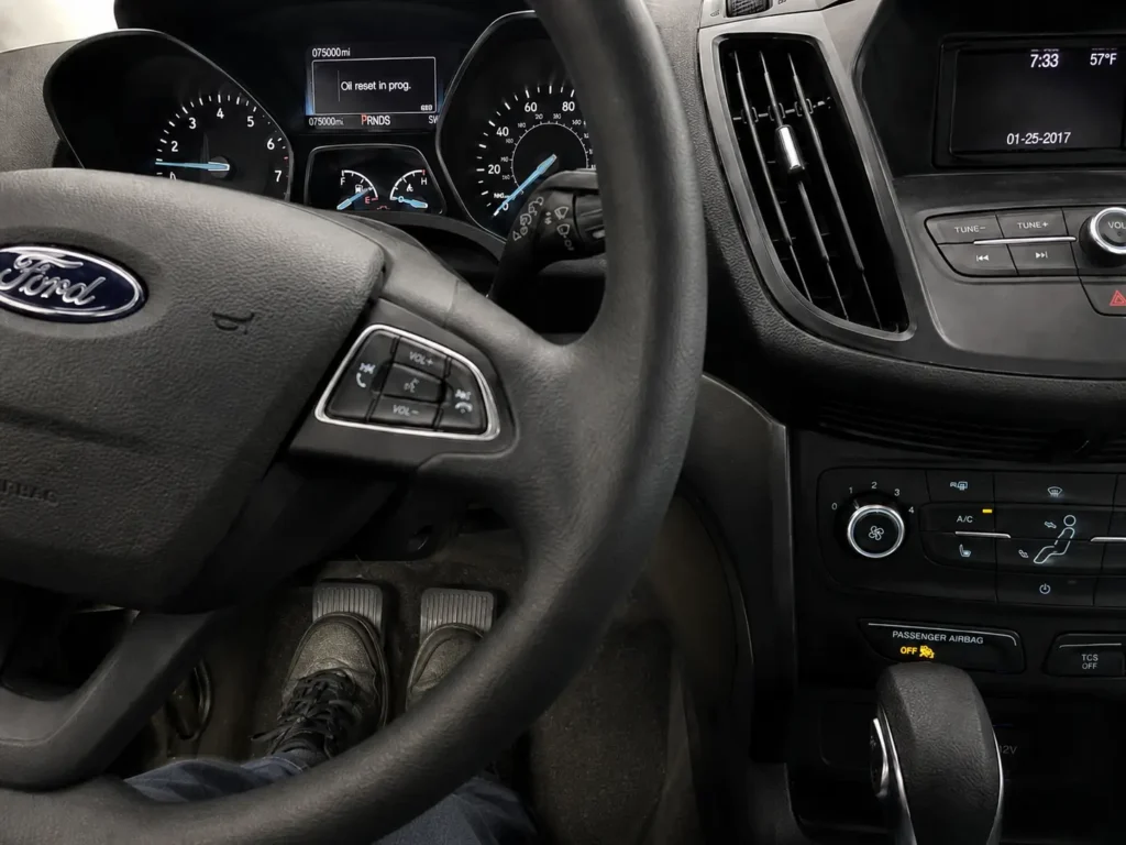 Inside a 2017 Ford Escape showing a driver pressing the accelerator and brake pedals while the dashboard displays “Oil reset in prog.