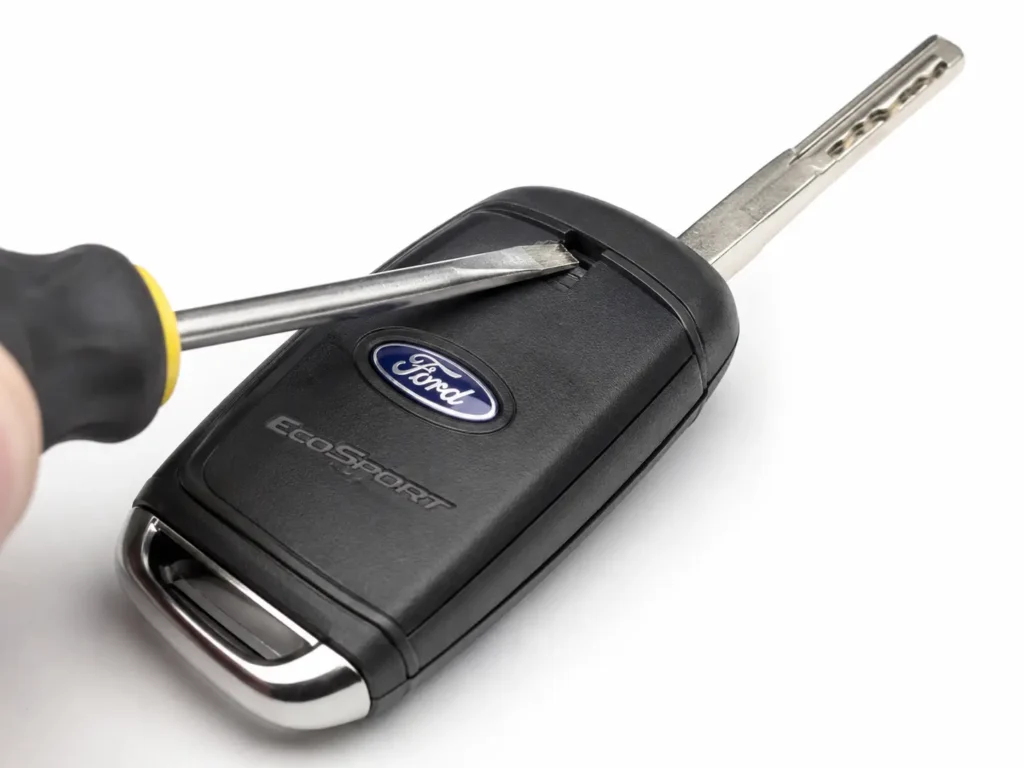 Close-up of a Ford EcoSport flip key with the switchblade key deployed while a flathead screwdriver is inserted into the notch near the blade cavity to open the battery cover.
