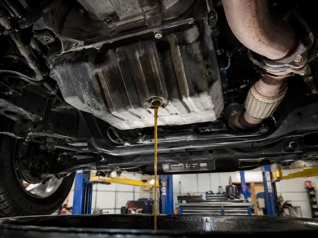 Underside of a Ford F-150 showing engine oil draining from the oil pan into a collection drain pan.